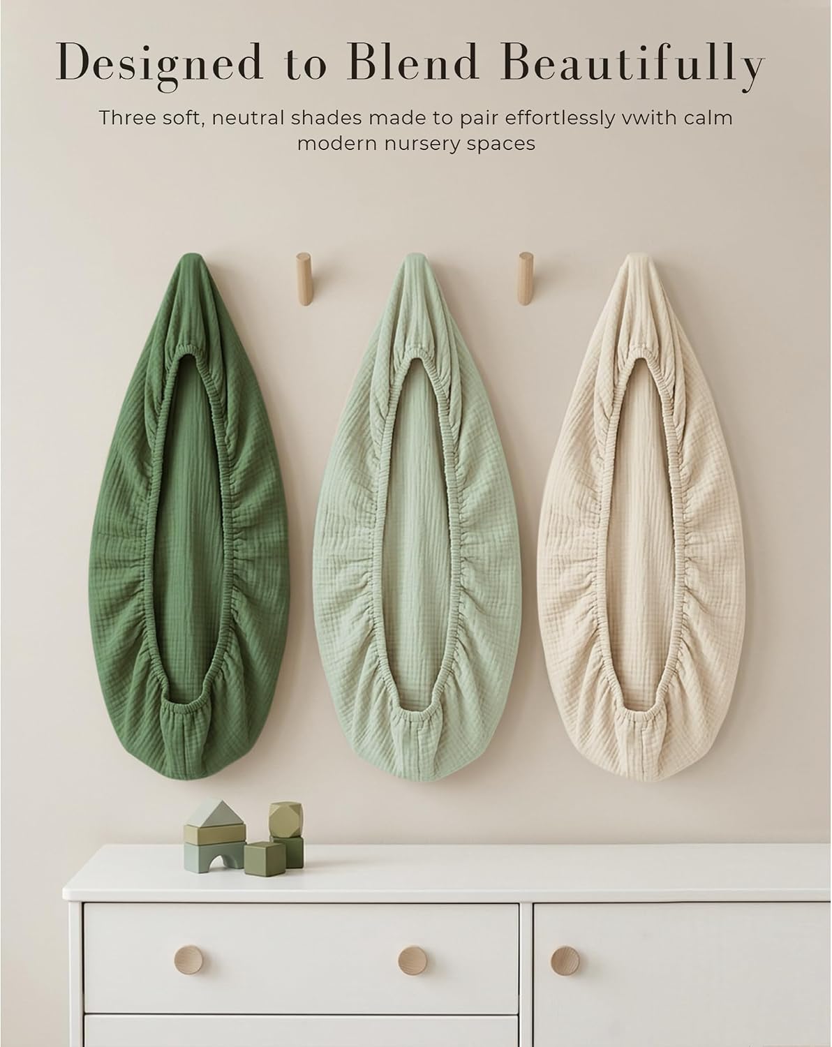 Three muslin bassinet fitted sheets in dark green, sage green, and cream, hung on wooden wall pegs above a white dresser. Text reads 'Designed to Blend Beautifully' with a subtitle about neutral nursery shades.