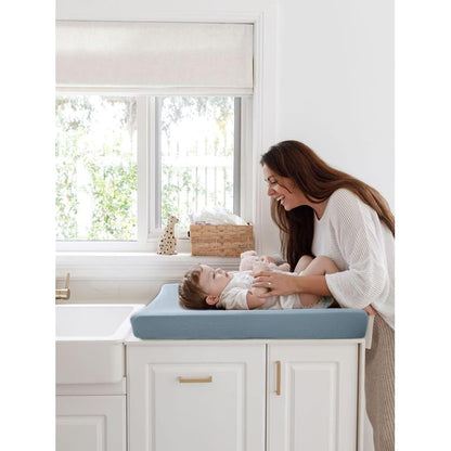 A smiling woman leans over a baby lying on a changing pad with a soft blue-gray cover. The pad sits on white cabinetry near a window. The cover has a subtle textured pattern.