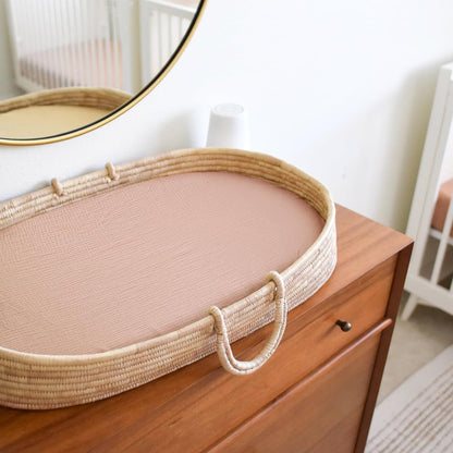 Oval woven rattan changing basket with a fitted dusty pink muslin sheet lining the interior, sitting on a medium-toned wood dresser in a nursery setting. A round mirror and white crib are visible in the background.