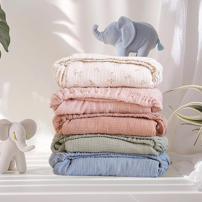 Stack of four folded muslin fitted crib sheets in white with small floral print, dusty pink, sage green, and blue. Two small stuffed elephant toys are placed beside and on top of the stack.