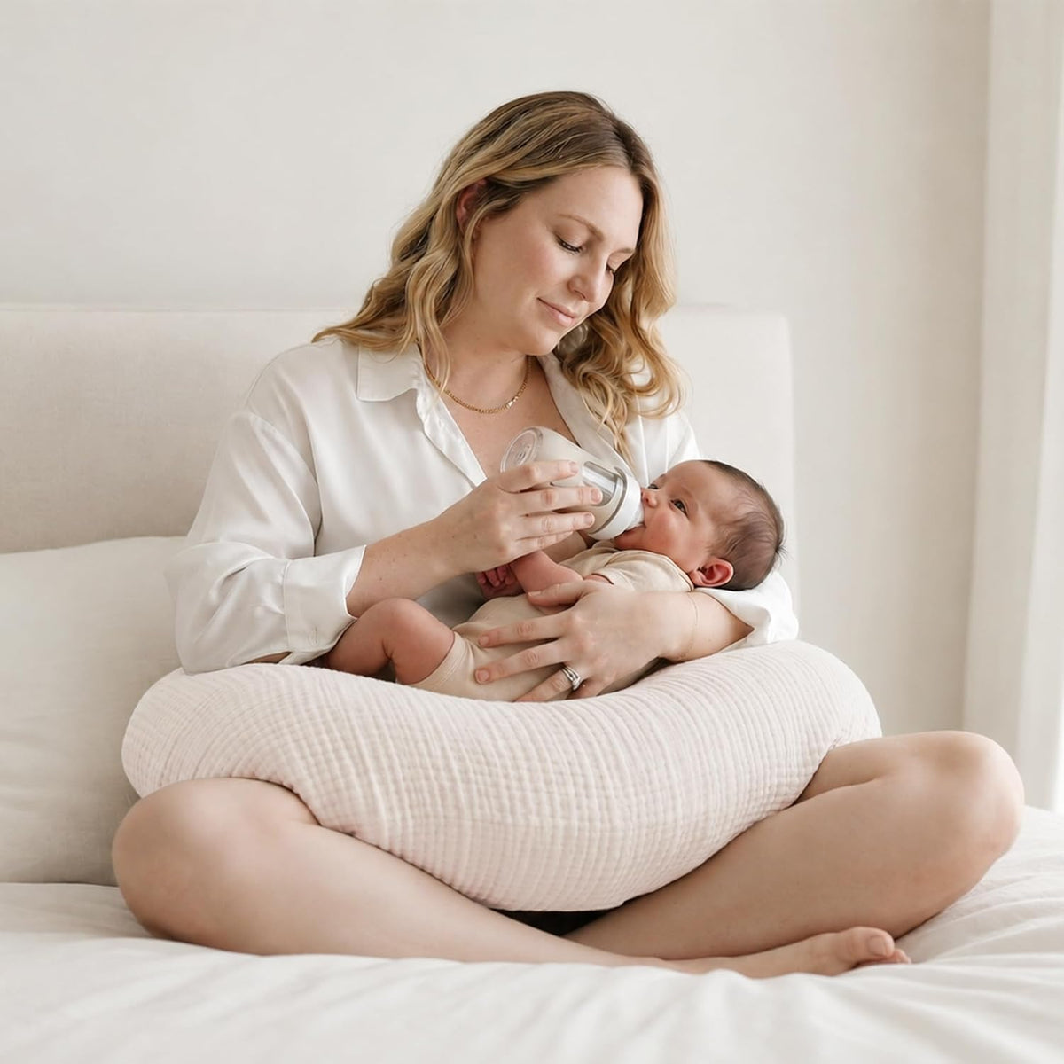 Woman sitting cross-legged on a white bed, bottle-feeding a newborn resting on a light pink textured nursing pillow in her lap. The pillow has a soft, ribbed cotton cover in a pale blush tone.