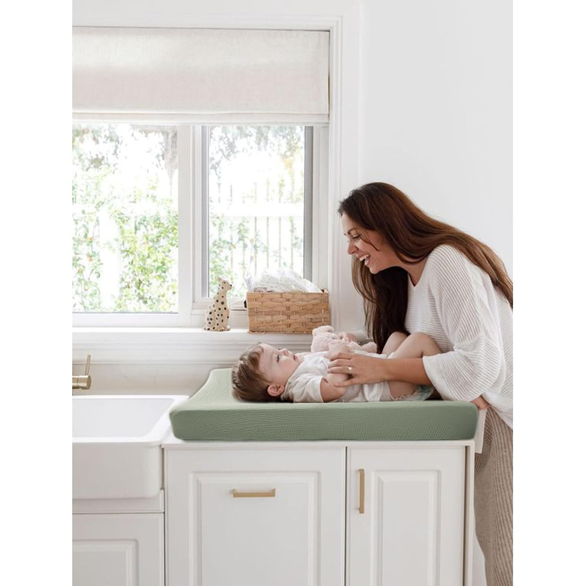 A smiling woman leans over a baby lying on a sage green changing pad cover atop white cabinetry. The textured, ribbed sage green cover has raised edges. A wicker basket and small decor sit on the windowsill behind.