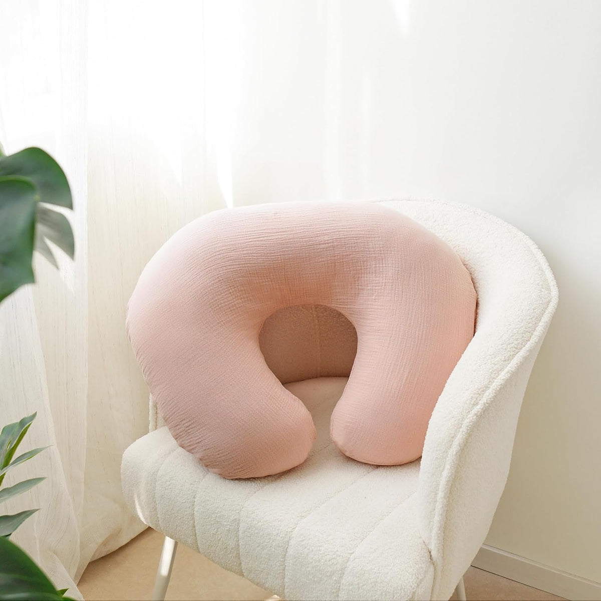 U-shaped nursing pillow in a soft pink muslin fabric cover, resting on a white textured armchair. The cover has a subtle woven texture and a rounded horseshoe silhouette, shown in a bright, airy room setting.