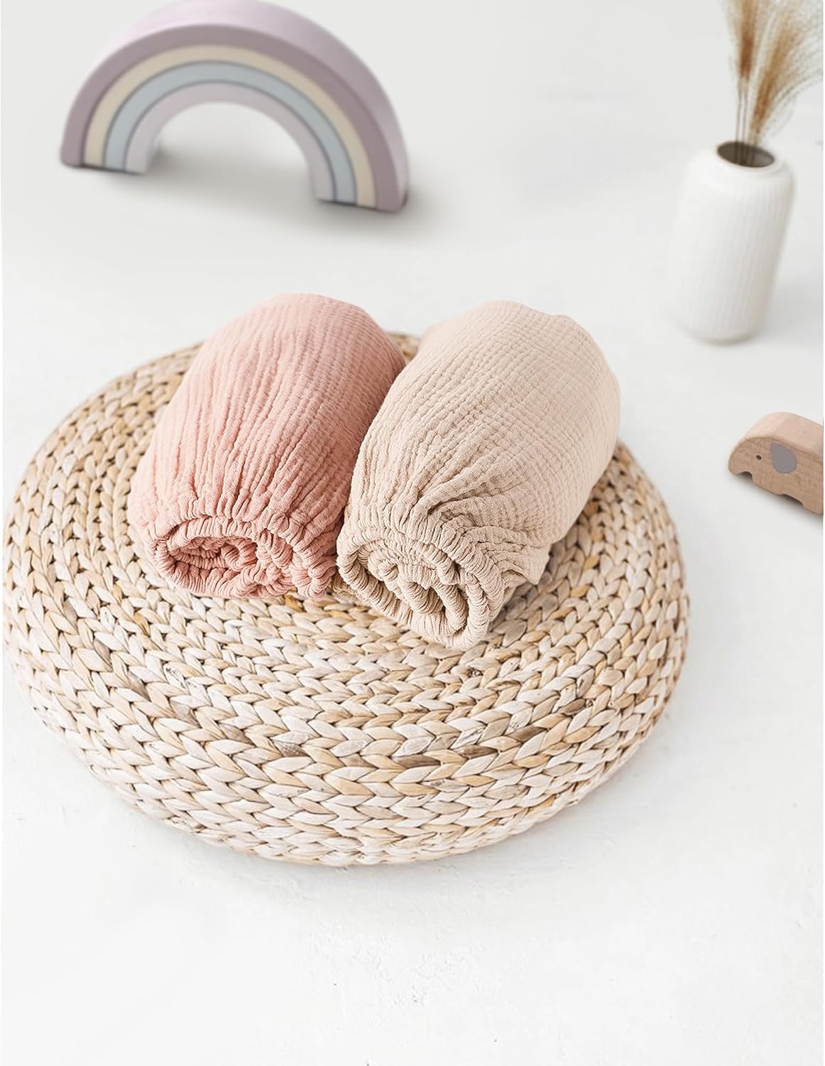Two rolled muslin fitted sheets in dusty pink and light beige, displayed on a round woven rattan trivet. Soft, textured cotton fabric with visible crinkled weave. A pastel rainbow toy and small white vase appear in the background.