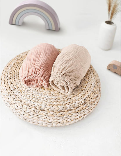 Two rolled muslin fitted sheets in dusty pink and light beige, displayed on a round woven rattan trivet. Soft, textured cotton fabric with visible crinkled weave. A pastel rainbow toy and small white vase appear in the background.
