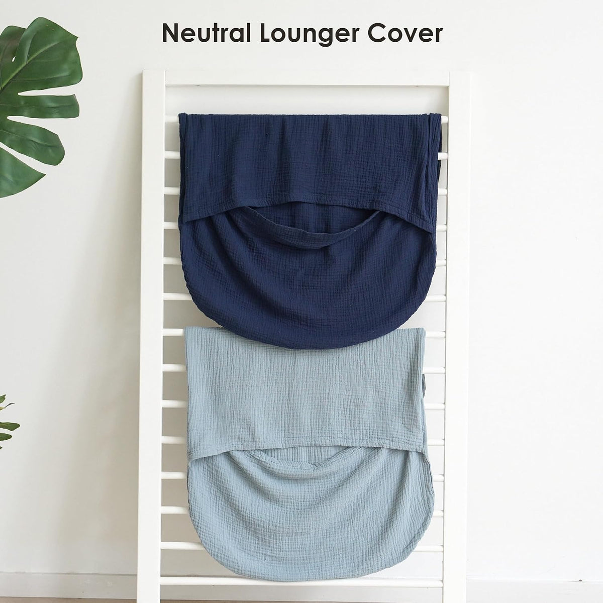 Two muslin baby lounger covers draped over a white ladder rack. The top cover is dark navy blue and the bottom is light dusty blue, both showing a textured woven fabric with a curved pouch shape. Text reads 'Neutral Lounger Cover'.