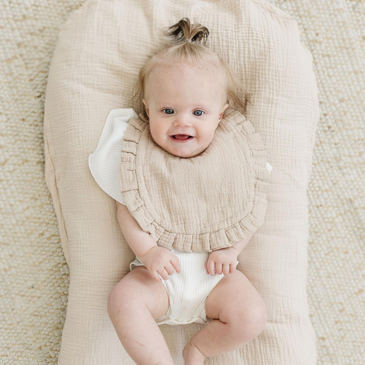 Smiling baby lying on a beige muslin lounger wearing a sandy beige muslin bib with ruffled edges over a white outfit. The bib has a soft, textured double-gauze fabric and a wide coverage design.