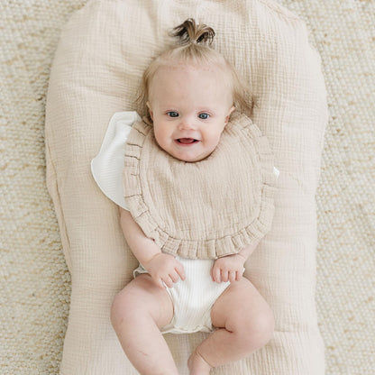 Smiling baby lying on a beige muslin lounger wearing a sandy beige muslin bib with ruffled edges over a white outfit. The bib has a soft, textured double-gauze fabric and a wide coverage design.