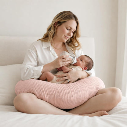 Woman sitting cross-legged on a white bed, bottle-feeding a newborn resting on a pink textured nursing pillow wrapped around her waist. The pillow has a soft, woven muslin-style cover in dusty pink.
