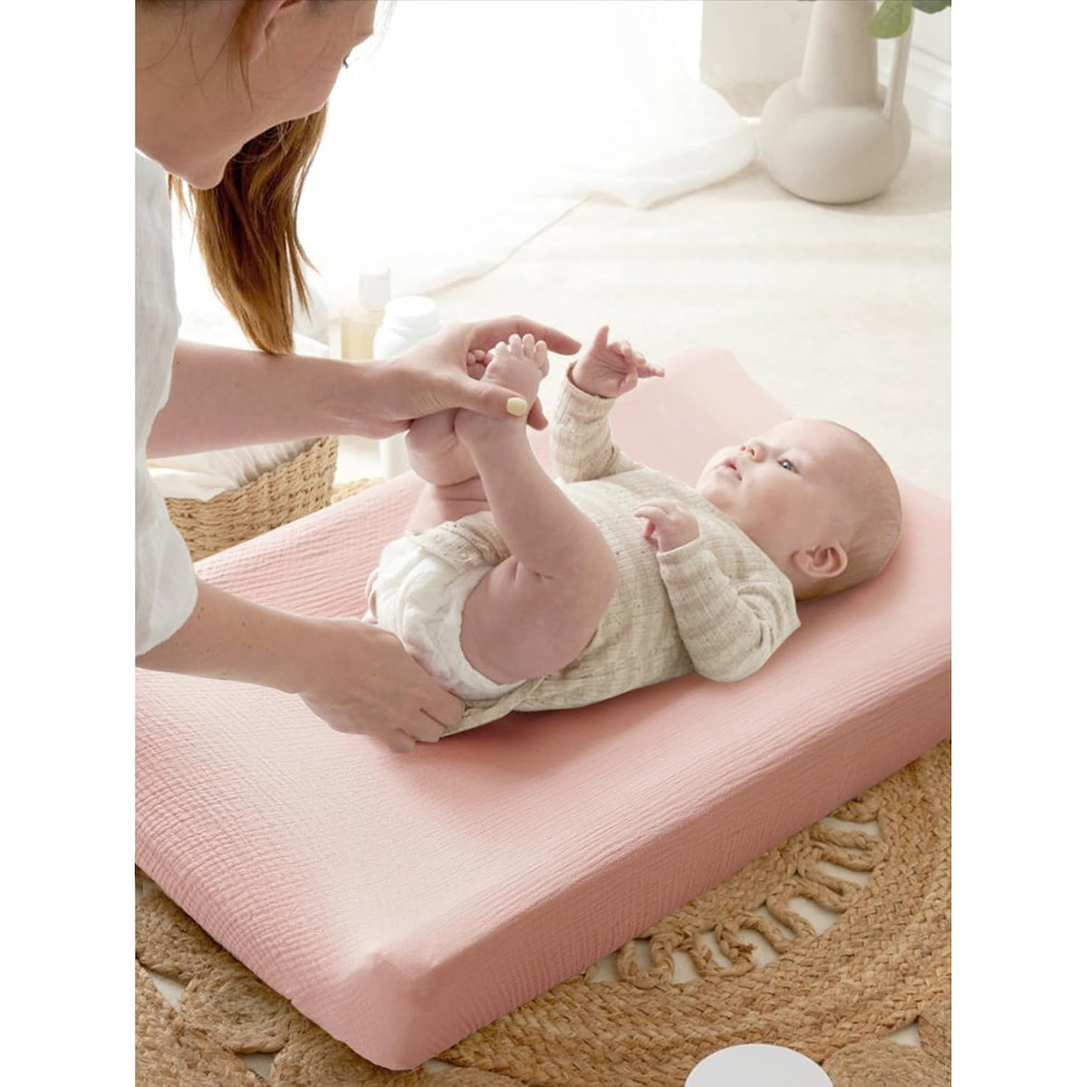 A baby lying on a dusty pink changing pad cover with a textured muslin-like surface. An adult holds the baby's legs during a diaper change. The cover fits snugly over a contoured changing pad.