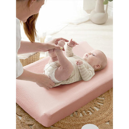 A baby lying on a dusty pink changing pad cover with a textured muslin-like surface. An adult holds the baby's legs during a diaper change. The cover fits snugly over a contoured changing pad.
