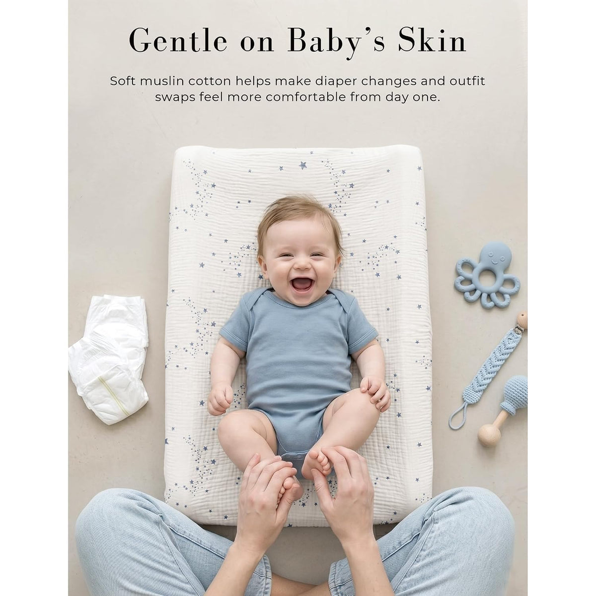 Baby lying on a changing pad covered with a white muslin cover featuring a blue star pattern, smiling up at a caregiver. A diaper and baby toys are visible nearby. Text reads 'Gentle on Baby's Skin.'