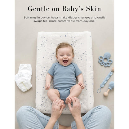 Baby lying on a changing pad covered with a white muslin cover featuring a blue star pattern, smiling up at a caregiver. A diaper and baby toys are visible nearby. Text reads 'Gentle on Baby's Skin.'