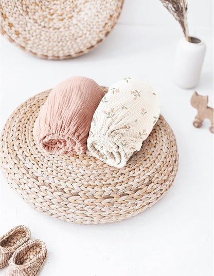 Two rolled muslin fitted bassinet sheets resting on a round woven rattan placemat. One sheet is solid dusty rose pink, the other is cream with a small floral print. Baby booties and decor visible nearby.