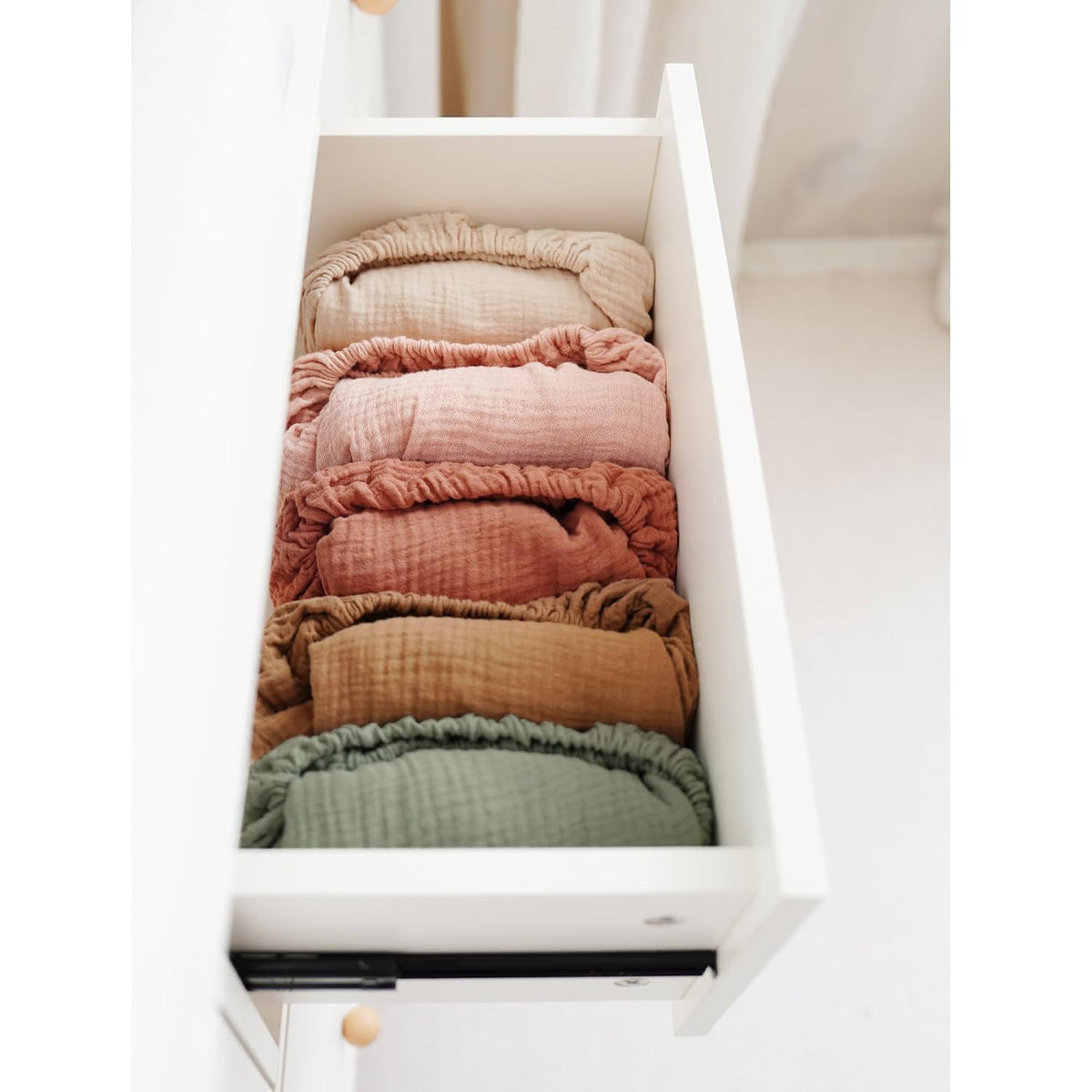 An open white drawer neatly stacked with six rolled muslin changing pad covers in a gradient of colors: beige, dusty rose, light pink, terracotta, tan, and sage green. Each cover shows visible elastic edges and textured muslin fabric.