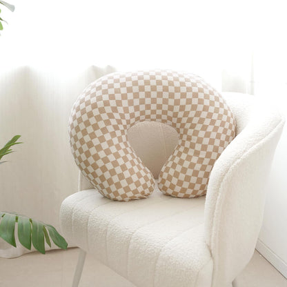 U-shaped nursing pillow cover in a tan and white checkerboard pattern, shown fitted on a nursing pillow resting on a white boucle chair. Smooth fabric with a small-scale checker print.