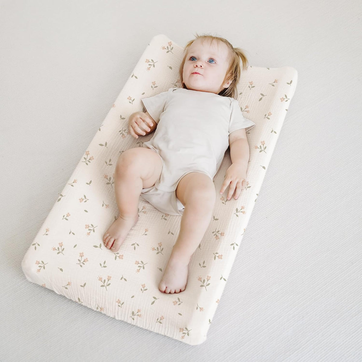 A baby lying on a rectangular changing pad covered in a cream muslin fabric with a small pink floral and green leaf print. The cover has a soft, textured surface and fitted edges wrapping the contoured pad.