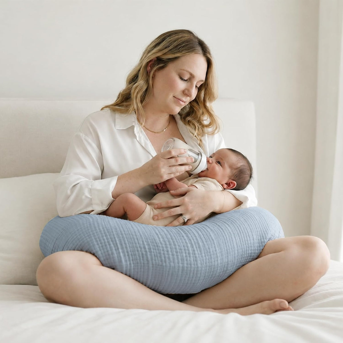Person sitting cross-legged on a white bed, bottle-feeding a newborn resting on a light blue textured nursing pillow. The pillow wraps around the lap and has a soft, ribbed muslin-like fabric in a muted blue tone.