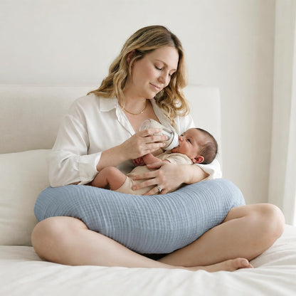 Person sitting cross-legged on a white bed, bottle-feeding a newborn resting on a light blue textured nursing pillow. The pillow wraps around the lap and has a soft, ribbed muslin-like fabric in a muted blue tone.