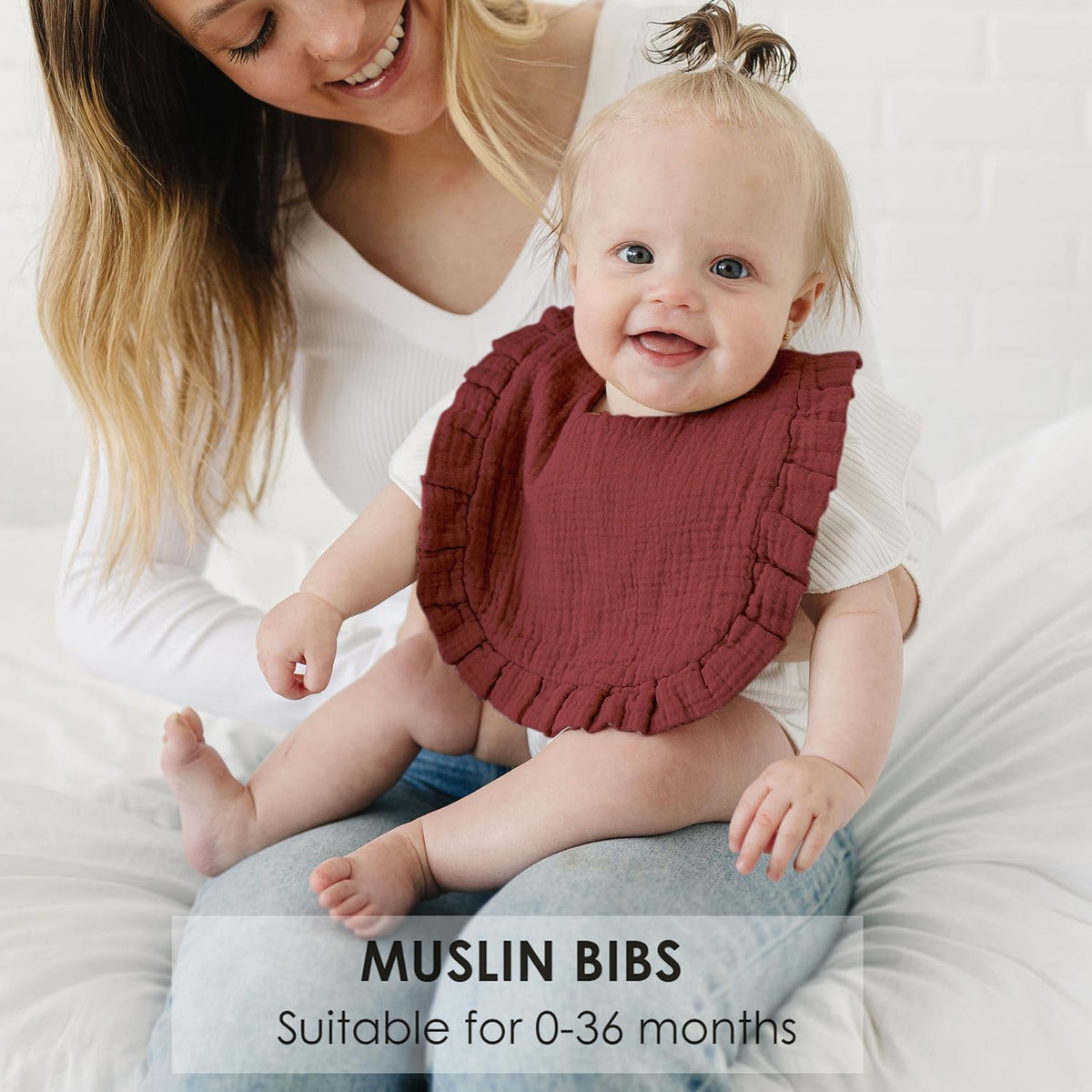 Smiling baby wearing a dark red muslin bib with ruffled edges, seated on an adult's lap on a white surface. Text reads 'MUSLIN BIBS - Suitable for 0-36 months.'