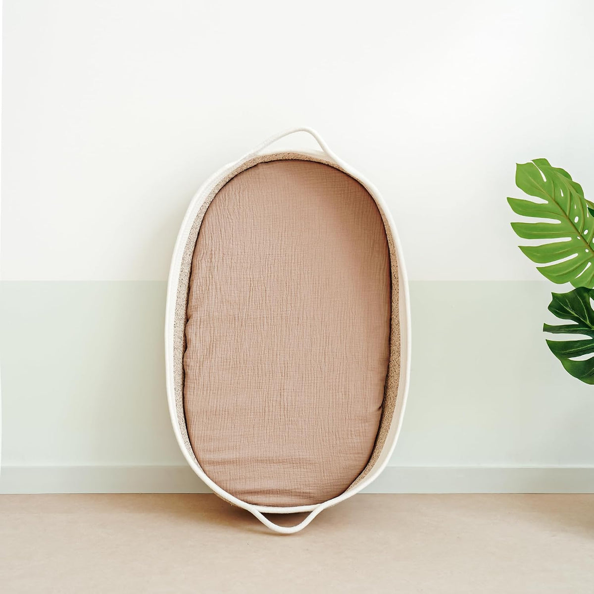 Oval bassinet with a dusty pink muslin fitted sheet lining its interior, leaned upright against a two-tone white and sage green wall. A monstera leaf is partially visible at right.