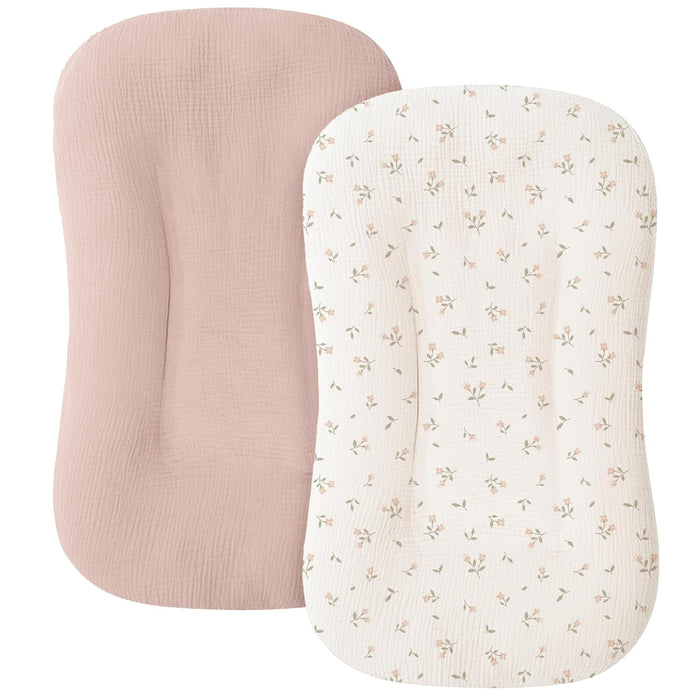 Two muslin baby lounger covers shown overlapping on a white background. The back cover is solid dusty pink; the front cover is cream with a small scattered floral print in pink and sage green.