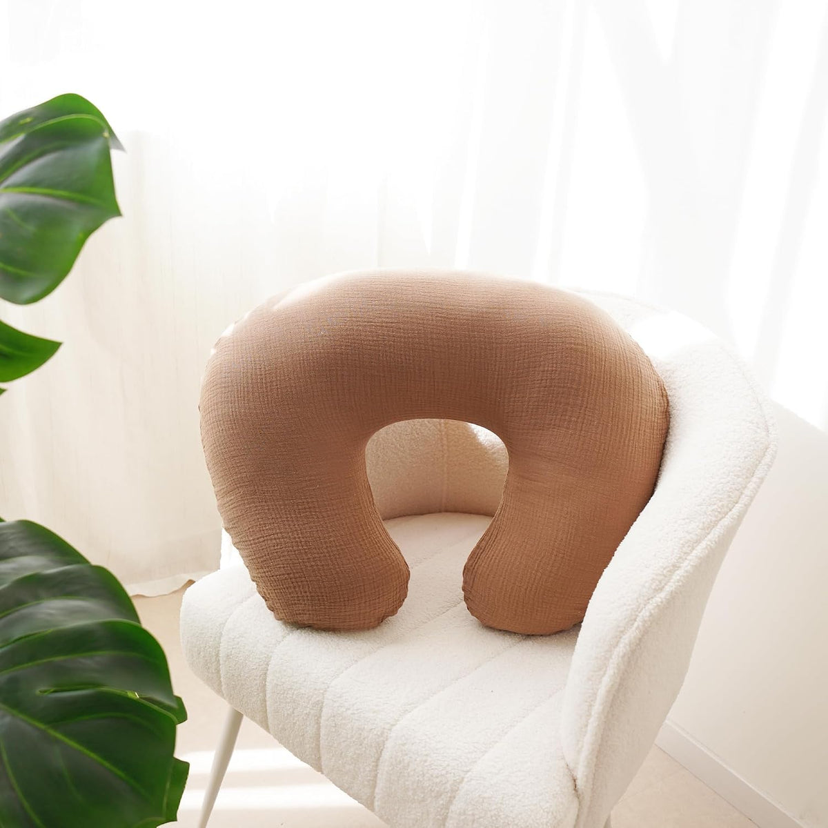 U-shaped nursing pillow with a warm tan/caramel textured muslin cover, resting on a white fluffy chair. The woven cotton fabric has a subtle grid-like texture. A monstera plant is visible in the background.