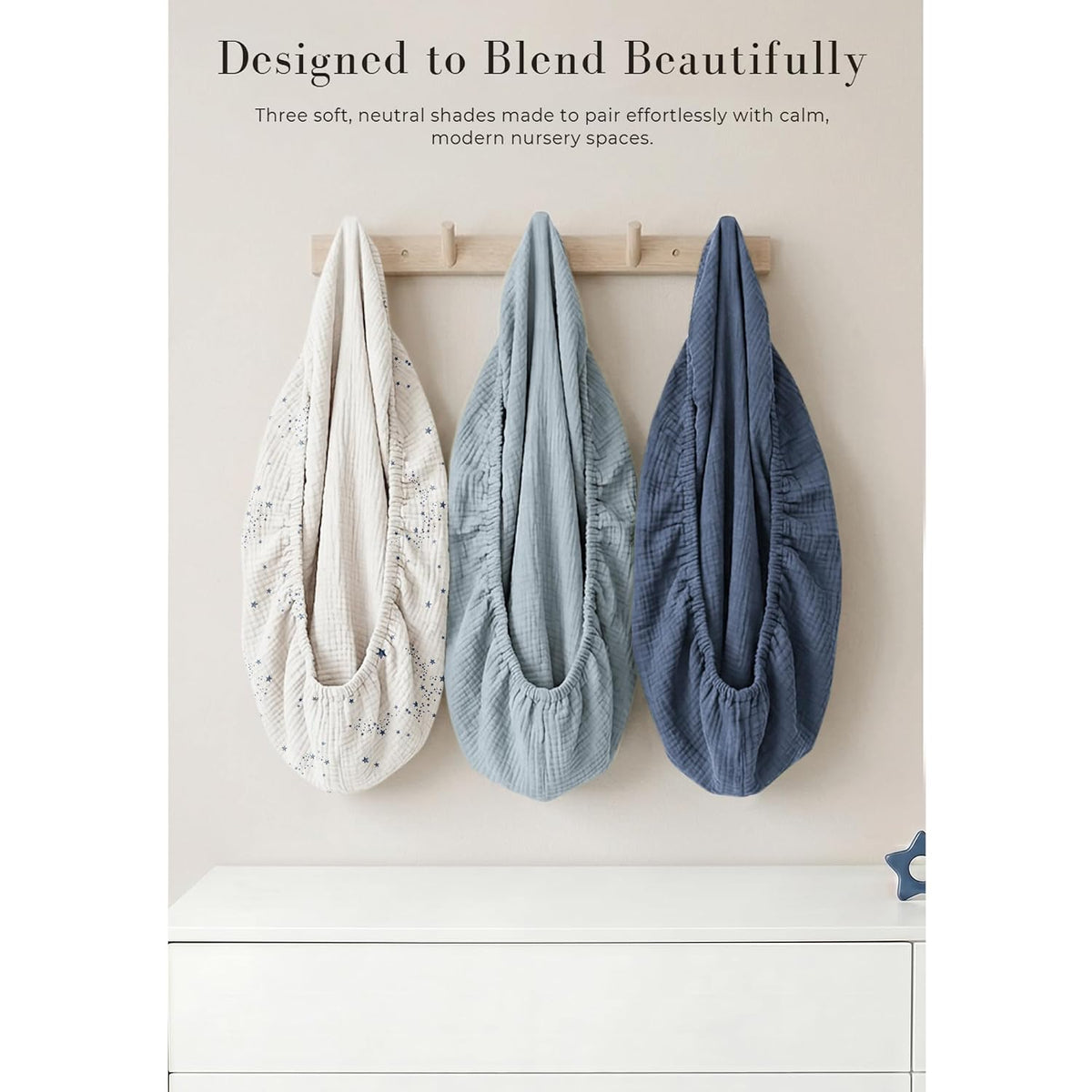 Three muslin changing pad covers hanging on a wooden wall hook rack. Left cover is cream with a small star/constellation print, center is light blue-gray, and right is medium blue, all showing gathered elastic edges and textured fabric.