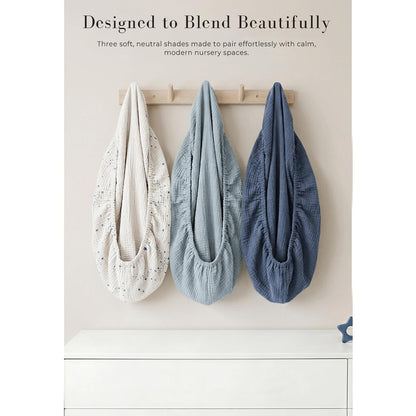 Three muslin changing pad covers hanging on a wooden wall hook rack. Left cover is cream with a small star/constellation print, center is light blue-gray, and right is medium blue, all showing gathered elastic edges and textured fabric.