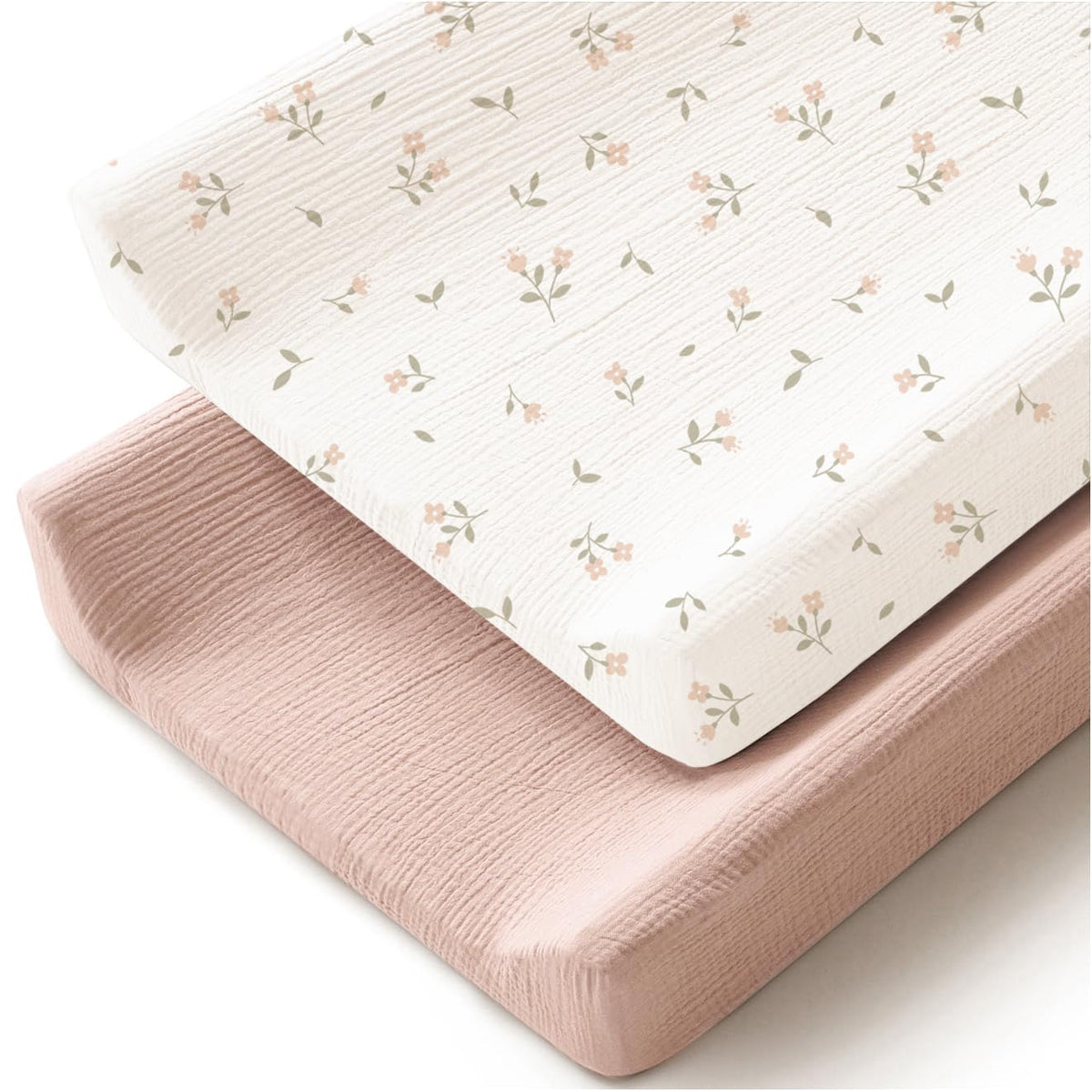Two changing pad covers stacked at an angle: one solid dusty pink and one white with a small scattered floral print in pink blooms and sage green leaves. Both have a textured muslin weave appearance.