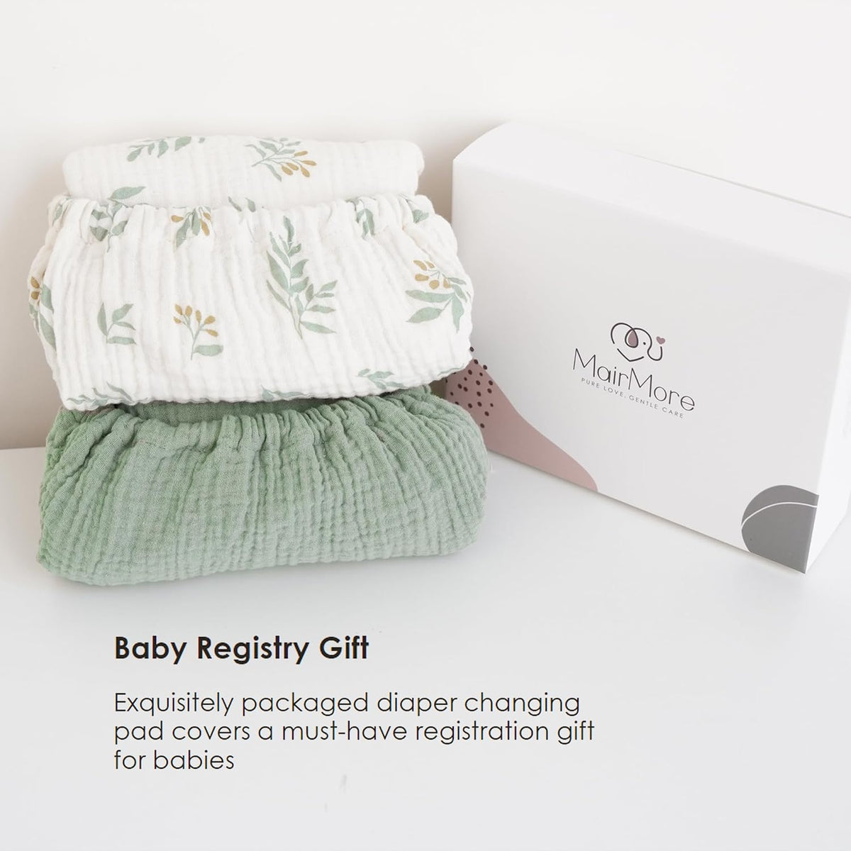 Two folded muslin changing pad covers stacked beside a white MairMore gift box. Top cover is white with a green and gold botanical leaf print; bottom cover is solid sage green. Textured cotton fabric visible.