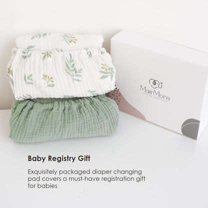 Two folded muslin changing pad covers stacked beside a white MairMore gift box. Top cover is white with a green and gold botanical leaf print; bottom cover is solid sage green. Textured cotton fabric visible.