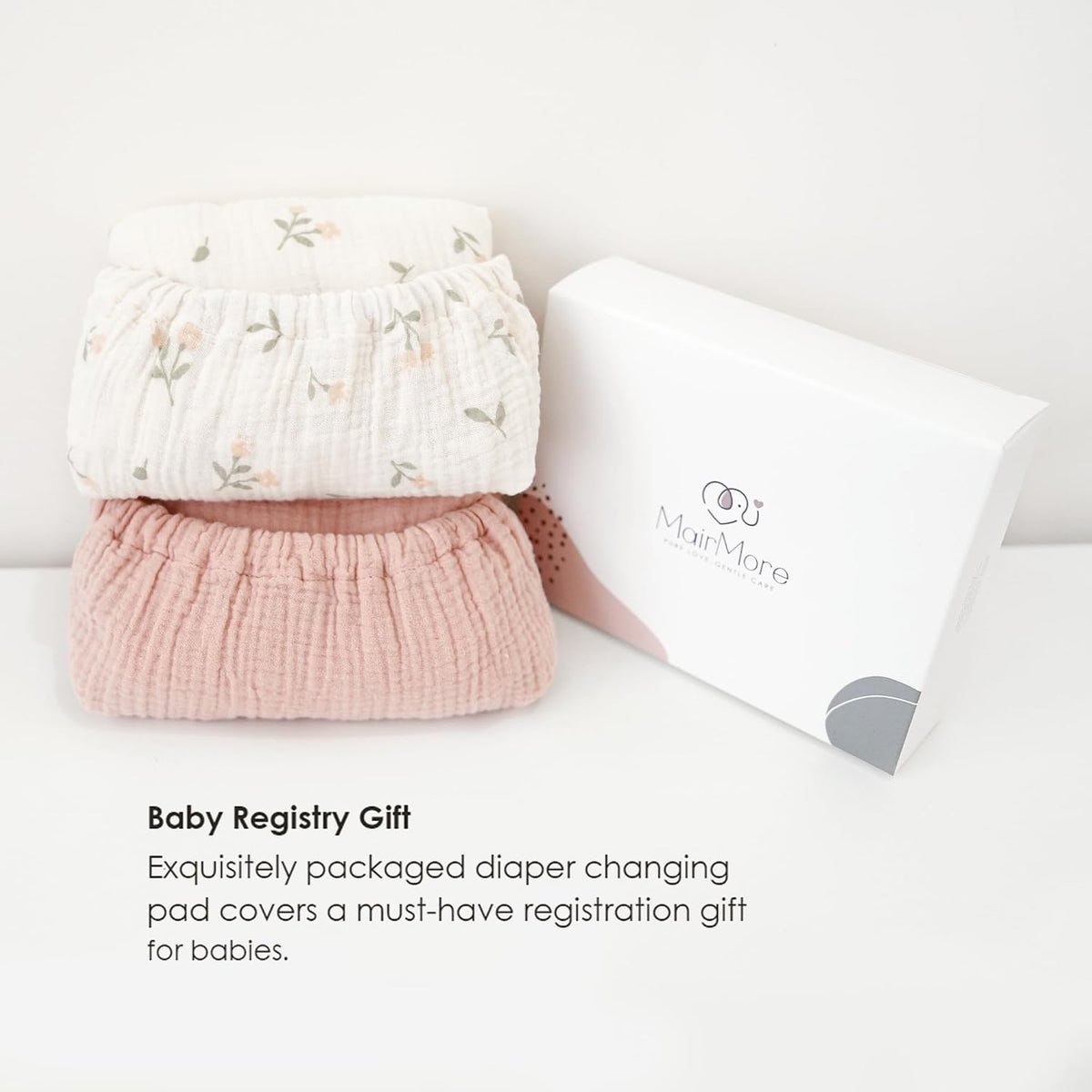 Two folded muslin changing pad covers stacked beside a white gift box branded MairMore. Top cover is white with a small floral print; bottom is solid dusty pink. Text below reads 'Baby Registry Gift.'