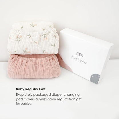 Two folded muslin changing pad covers stacked beside a white gift box branded MairMore. Top cover is white with a small floral print; bottom is solid dusty pink. Text below reads 'Baby Registry Gift.'