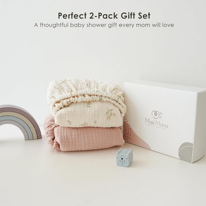 Two folded muslin fitted bassinet sheets stacked together: a cream sheet with small floral print on top and a solid dusty pink sheet below. Shown beside a white MairMore gift box, a small rainbow toy, and a tiny bear cube.