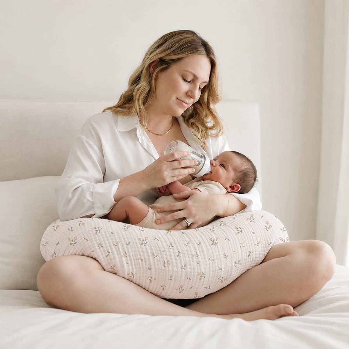 Woman seated cross-legged on a bed bottle-feeding a newborn resting on a crescent-shaped nursing pillow. The pillow cover is light pink with a small floral print and a textured muslin-style fabric.