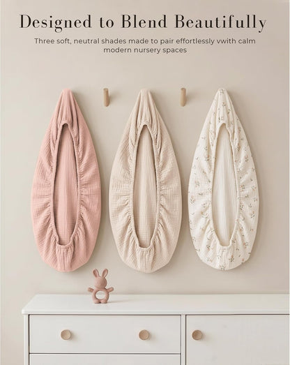 Three fitted bassinet sheets displayed on a wall in pink, beige, and cream floral muslin. Text reads 'Designed to Blend Beautifully' above. A small dresser with a bunny toy sits below.
