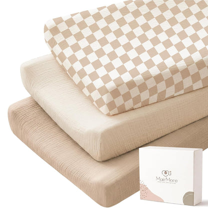 Three stacked changing pad covers in muslin fabric: top cover has a tan and cream checkerboard pattern, middle is solid cream, bottom is solid tan. A white MairMore gift box is shown in the foreground.