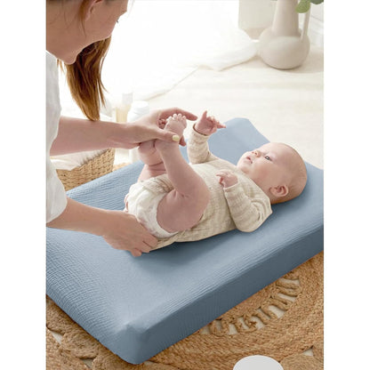 A baby lying on a changing pad covered in a soft blue textured muslin cover, while an adult holds the baby's legs. The cover has a subtle waffle-like texture and fits snugly over the contoured pad.