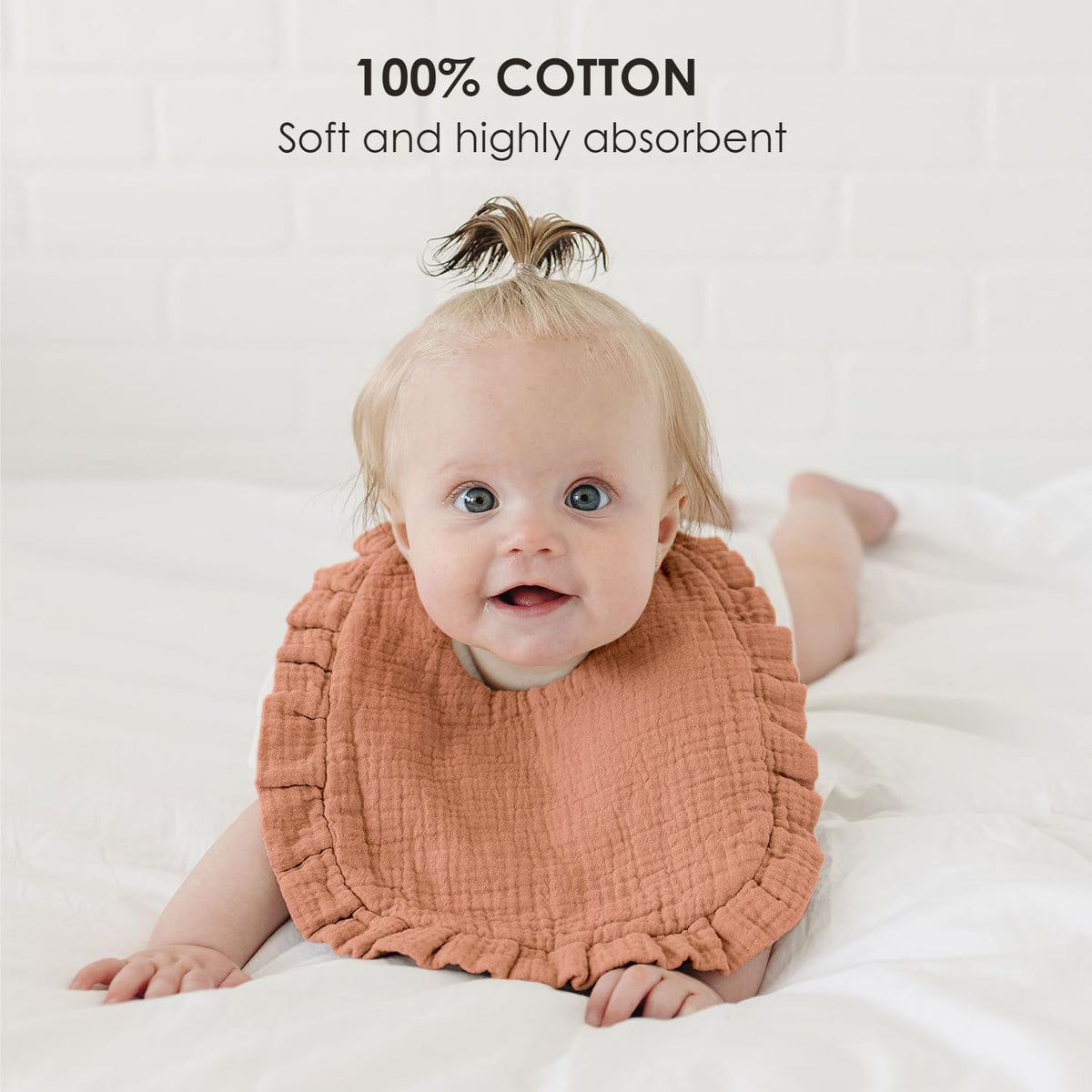 Smiling baby wearing a terracotta-colored muslin bib with ruffled edges, lying on a white surface. Text overlay reads '100% COTTON - Soft and highly absorbent.'