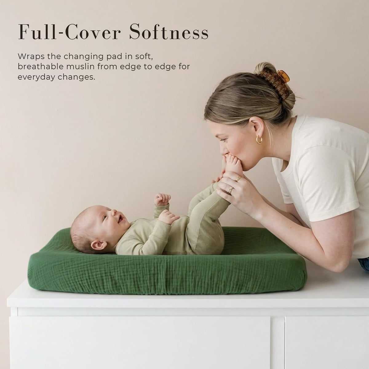 A baby lies on a dark green muslin changing pad cover atop a white dresser while a woman kisses the baby's foot. Text reads 'Full-Cover Softness' with a description of breathable muslin coverage.