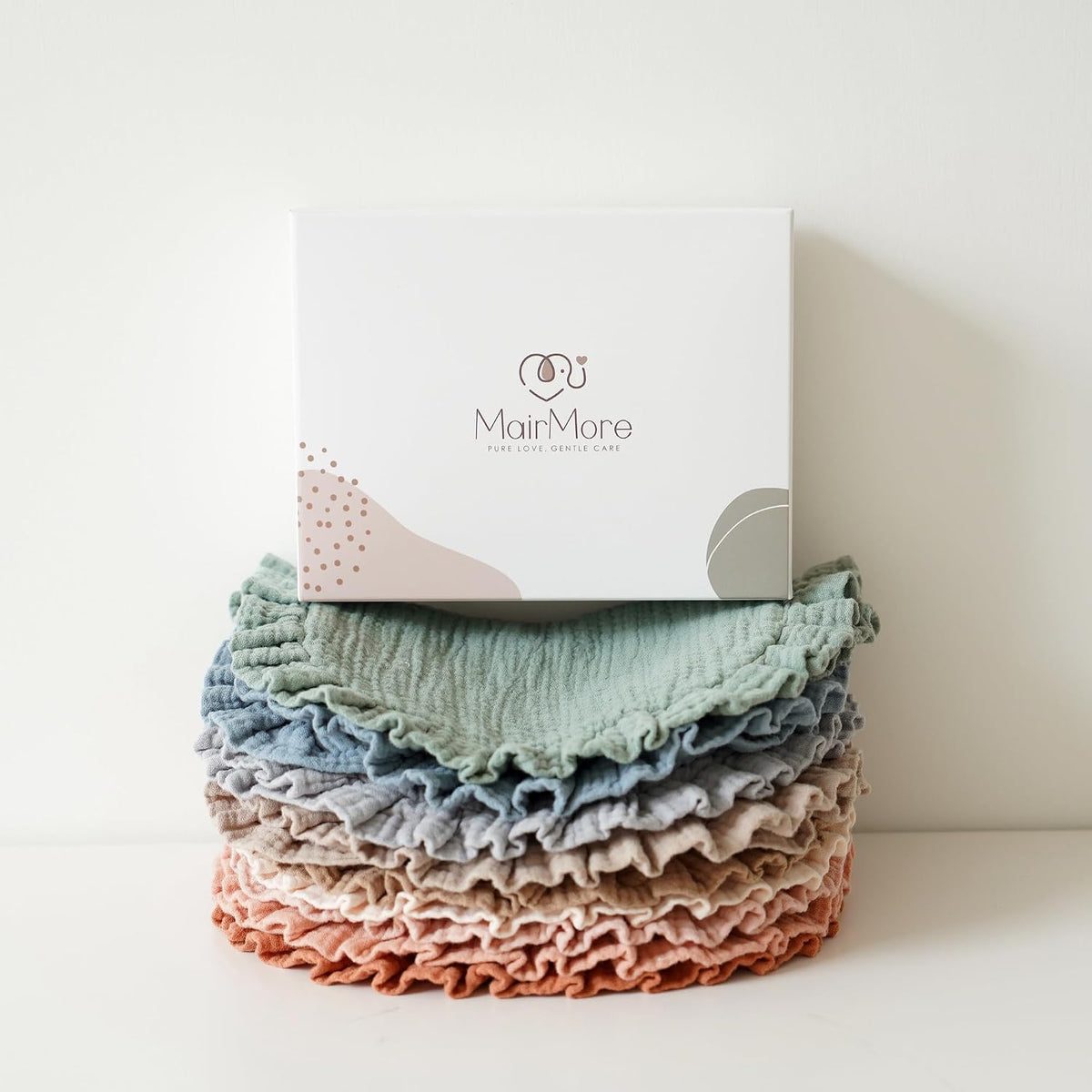 Stack of MairMore muslin bandana bibs in layered pastel colors -- sage green, blue, gray, blush, and terracotta -- with ruffled edges, topped by a white branded gift box reading 'MairMore, Pure Love, Gentle Care'.