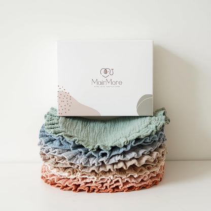 Stack of MairMore muslin bandana bibs in layered pastel colors -- sage green, blue, gray, blush, and terracotta -- with ruffled edges, topped by a white branded gift box reading 'MairMore, Pure Love, Gentle Care'.
