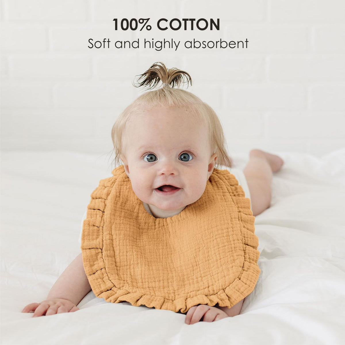 Baby wearing a golden-yellow muslin bib with ruffled edges, lying on a white surface. Text overlay reads '100% COTTON - Soft and highly absorbent.' The bib has a textured, crinkled fabric appearance.
