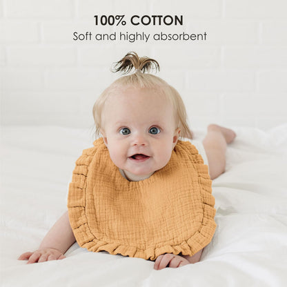 Baby wearing a golden-yellow muslin bib with ruffled edges, lying on a white surface. Text overlay reads '100% COTTON - Soft and highly absorbent.' The bib has a textured, crinkled fabric appearance.