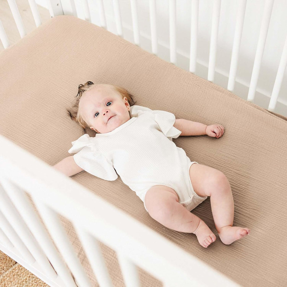 Baby lying on a tan muslin fitted crib sheet in a white crib, viewed from above. The sheet has a soft, textured weave and fits snugly over the mattress. Baby wears a white short-sleeve onesie.