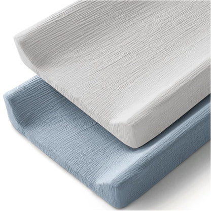 Two changing pad covers stacked at an angle on a white background. The top cover is white and the bottom is soft blue, both showing a textured muslin weave fabric that wraps snugly around rectangular foam pads.