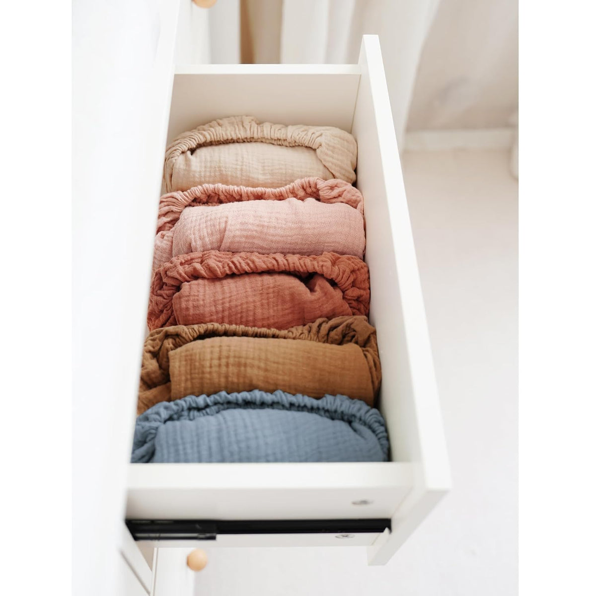 An open white dresser drawer holding six rolled muslin changing pad covers in a gradient of colors: tan, dusty rose, light pink, terracotta, golden brown, and muted blue. Textured crinkled fabric with elastic edges visible.