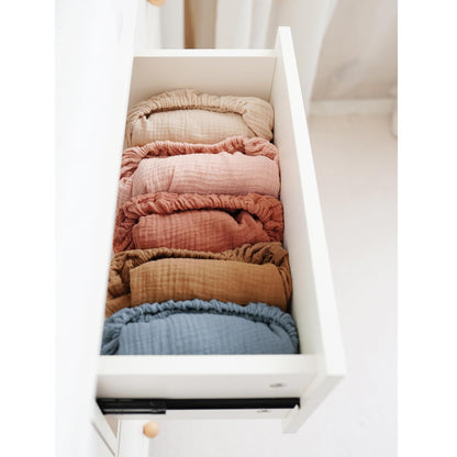 An open white dresser drawer holding six rolled muslin changing pad covers in a gradient of colors: tan, dusty rose, light pink, terracotta, golden brown, and muted blue. Textured crinkled fabric with elastic edges visible.