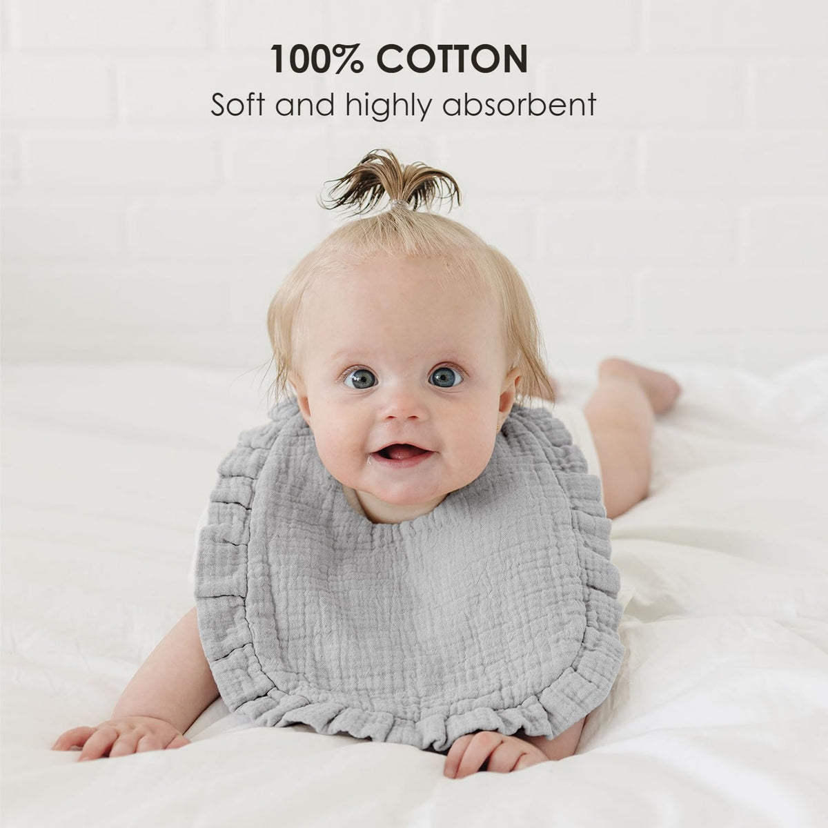 Smiling baby wearing a light gray muslin cotton bib with ruffled edges, lying on a white surface. Text overlay reads '100% COTTON - Soft and highly absorbent.'